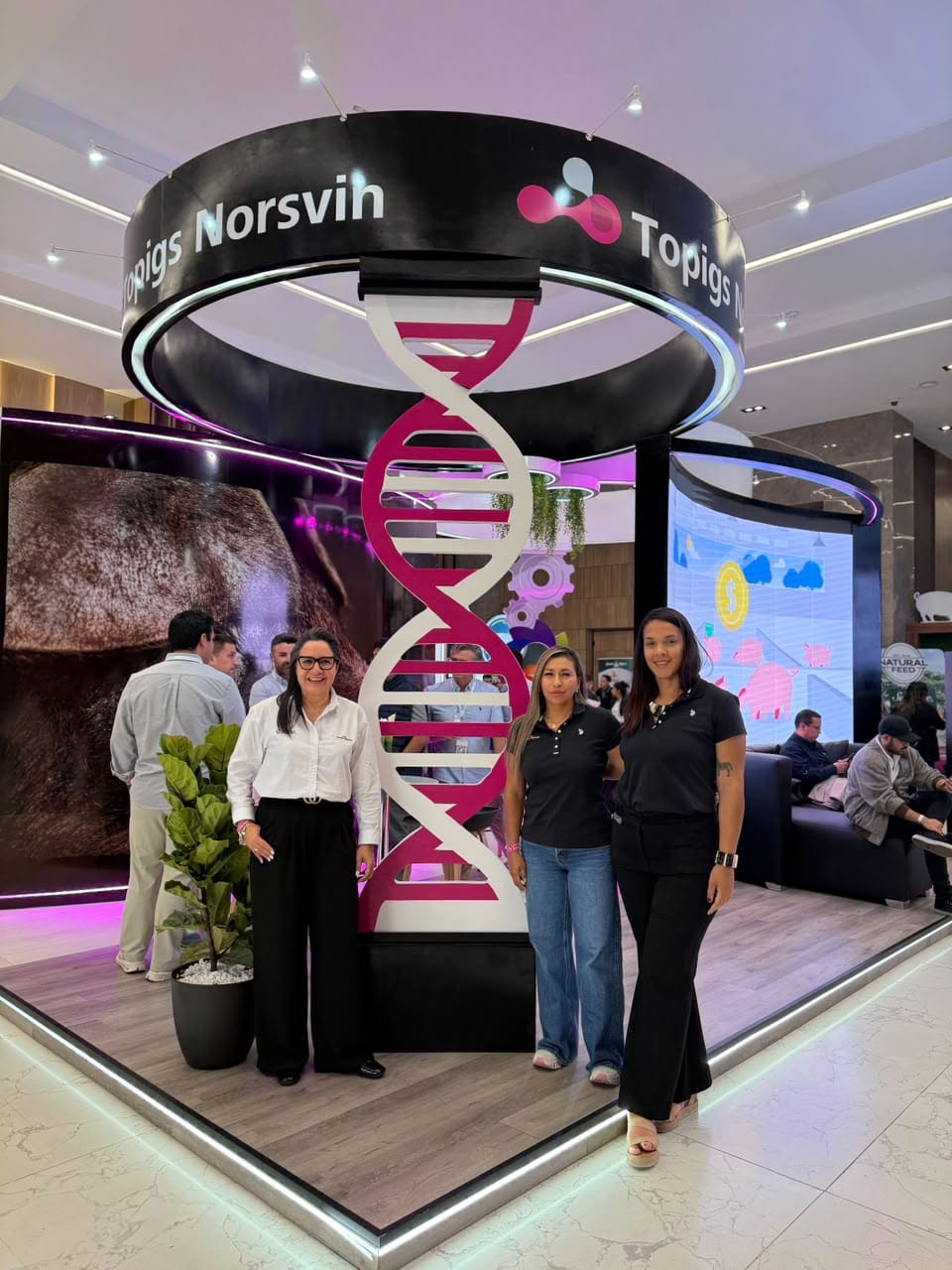 Three people pose in front of the Topigs Norsvin stand, beside a large DNA helix and below a ring bearing the brand logo
